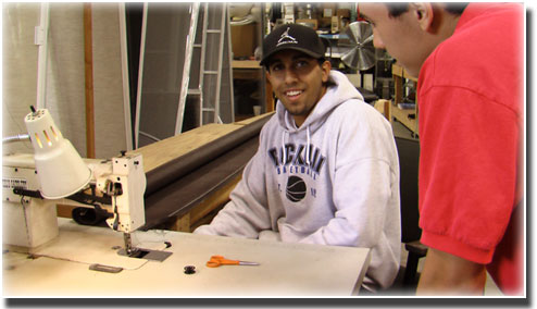 man in sweatshirt and black baseball hat sitting at a table with a sewing machine and scissors on it