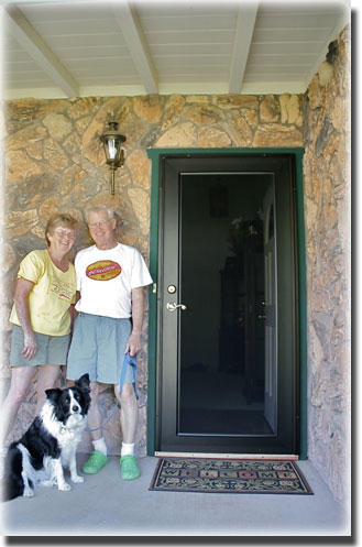 Eldery couple with dog standing by black security door on front porch doormat in front of door