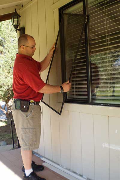 Man in shorts and red shirt installing window screen in window of home