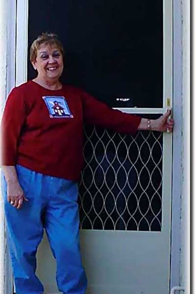 Middle aged woman in red shirt and jeans standing in front of her screen door at her home