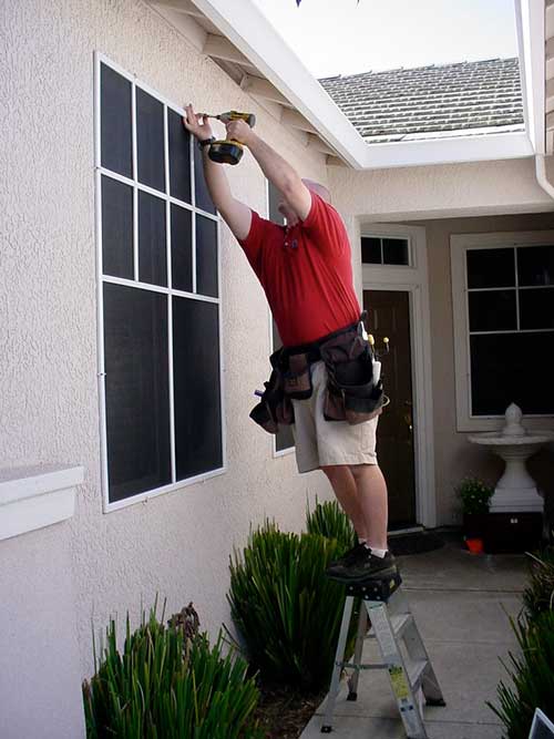 man in red shirt and shorts holding a drill while standing on a small ladder installing window screens