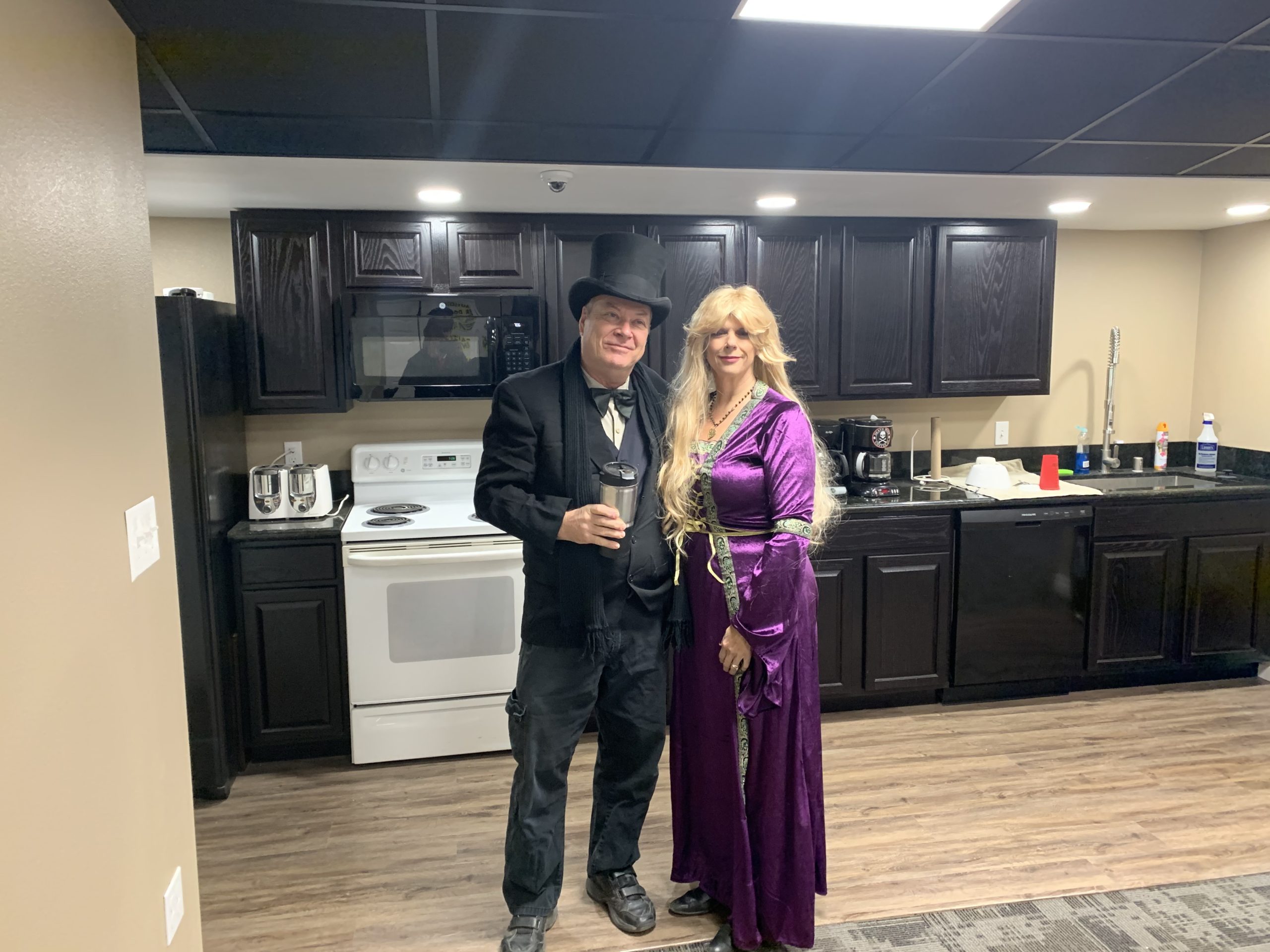 man dressed in black and blonde haired woman in purple standing in kitchen of a home